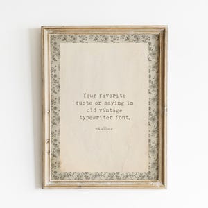 Può includere: Una cornice rettangolare in legno contenente un foglio color crema con il testo nero: Your favorite quote or saying in old vintage typewriter font. -Author. Il foglio è bordato da un passe-partout con motivo floreale grigio e bianco.
