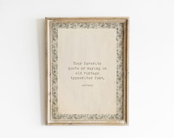 May include: A rectangular wooden frame containing a cream-coloured paper with the black text: Your favourite quote or saying in old vintage typewriter font. -Author. The paper is bordered by a grey and white floral patterned matting.