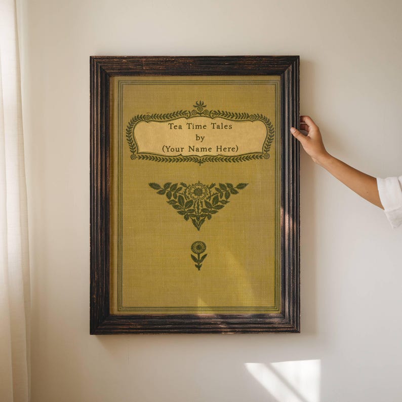 May include: A framed print of a book cover with a vintage design. The title is "Tea Time Tales" and the text below reads "by (Your Name Here)". The cover is a light yellow color with green floral designs.