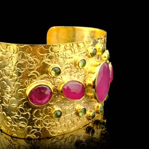 May include: A gold-colored cuff bracelet with a textured surface and a series of embedded gemstones. The bracelet features large, oval-shaped ruby-colored stones and smaller, round, green-colored stones.