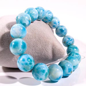 May include: A bracelet made of large, round, light blue beads with white marbling. The beads are strung on a clear elastic cord. The bracelet is displayed on a grey, textured surface. The beads have a polished, glossy finish.