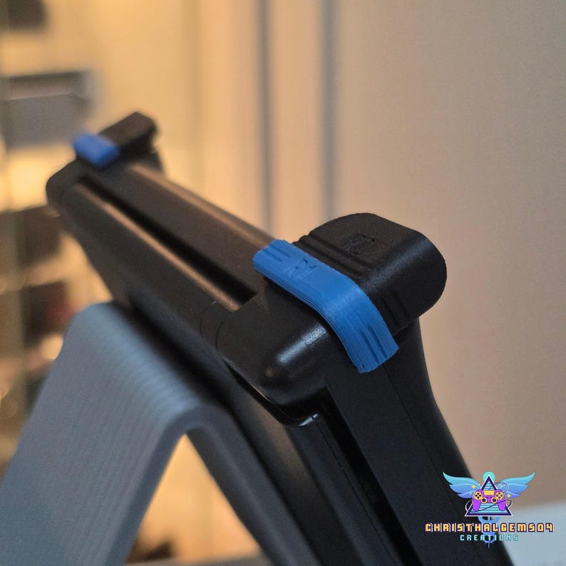 May include: A black handheld gaming console accessory featuring bright blue plastic clips attached to the top edges. The device is mounted on a light gray stand. The text "CHRISTHALGEM504 CREATIONS" is visible in the bottom right corner.