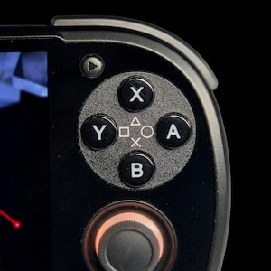 May include: Close-up of a black handheld gaming device. The device has a directional pad, X, Y, A, and B buttons, and a joystick. The buttons are grey with white lettering and symbols. The joystick has a red illuminated ring.