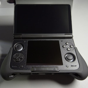 May include: Black handheld gaming console with a clamshell design. The console features two screens, a directional pad, buttons, and a joystick. The console has a matte finish and a grip for comfortable handling. The screen is black and the console is open.