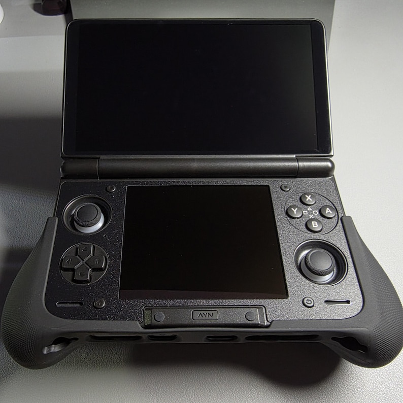 May include: Black handheld gaming console with a clamshell design. The console features two screens, a directional pad, buttons, and a joystick. The console has a matte finish and a grip for comfortable handling. The screen is black and the console is open.