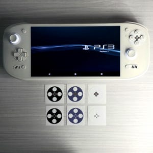 Può includere: Una console di gioco portatile bianca con uno schermo che mostra il logo PlayStation 3. Sei adesivi abbinati con disegni di pulsanti del controller sono sotto la console. La console ha pulsanti e joystick su entrambi i lati.