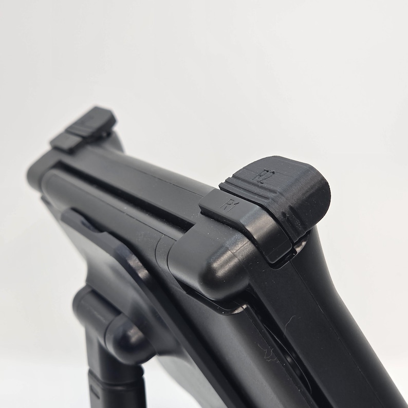 May include: A black gaming controller accessory featuring prominent R1 and R2 shoulder buttons. The device has a matte finish and a structured, ergonomic design with visible hinge components, focusing on the top trigger button layout.