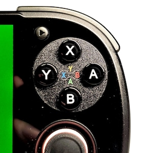 May include: Close-up of a black handheld gaming device. The device features a green screen and a circular button pad with X, Y, A, and B buttons. The buttons are black with white lettering. A play button is visible.
