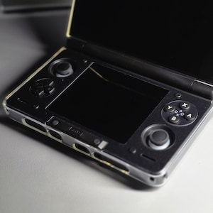 May include: A black handheld gaming console with a clear protective case. The console is open, revealing two screens. The bottom screen has a joystick and buttons. The top screen is black. The console is on a light gray surface.