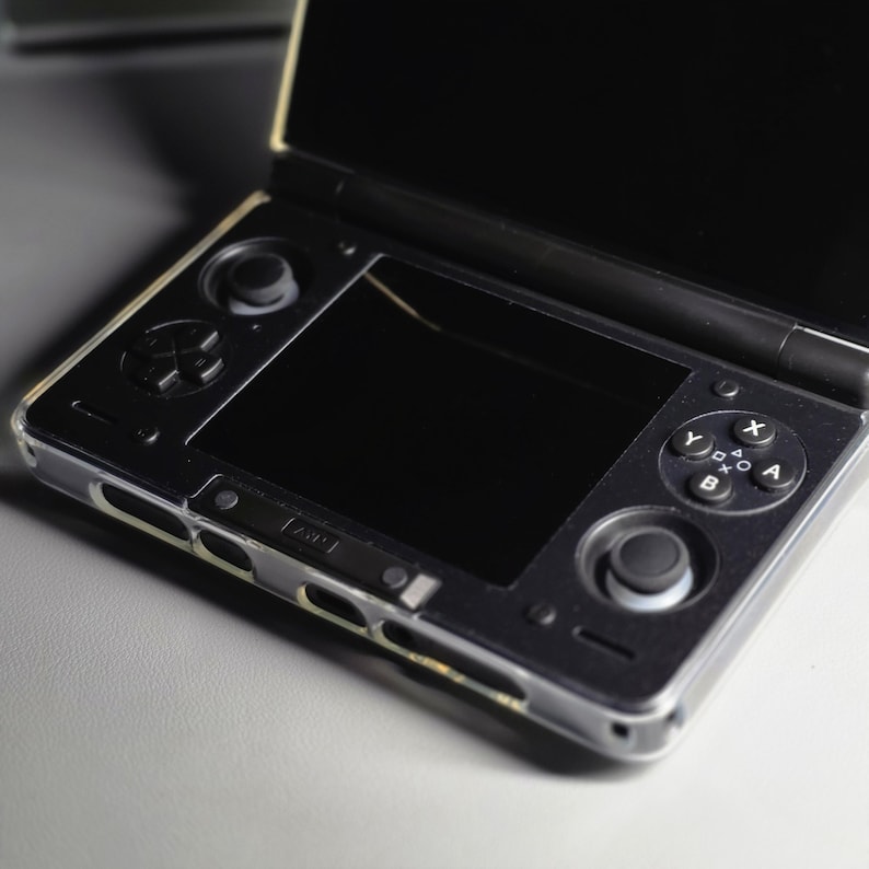 May include: A black handheld gaming console with a clear protective case. The console is open, revealing two screens. The bottom screen has a joystick and buttons. The top screen is black. The console is on a light gray surface.
