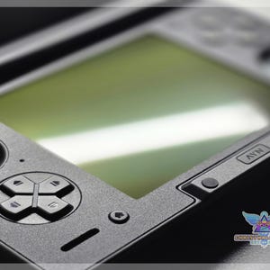 May include: Close-up of a black handheld gaming console. The device features a directional pad, buttons, and a screen with a green tint. The console has a textured surface and a logo in the bottom right corner.