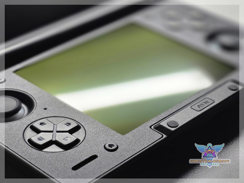 May include: Close-up of a black handheld gaming console. The device features a directional pad, buttons, and a screen with a green tint. The console has a textured surface and a logo in the bottom right corner.
