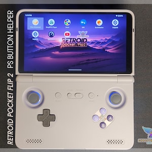 May include: A white Retroid Pocket Flip 2 handheld gaming device with a screen displaying app icons and a purple landscape. The device has a D-pad, buttons, and joysticks. Text on the device reads "RETROID pocket/Flip2".