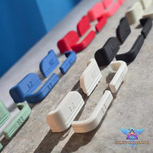May include: A collection of replacement controller shoulder buttons in various colors including red, blue, black, white, and light green. Each button is marked with L1, L2, R1, or R2 labels. The components are arranged in rows on a textured surface.
