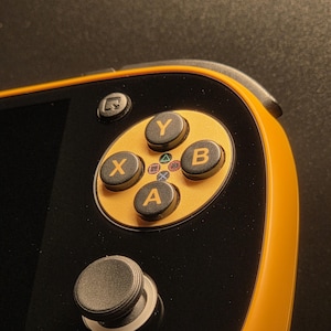 Peut inclure: Gros plan sur une console de jeu portable avec un corps noir et des accents orange. L'appareil comporte un pavé directionnel doré avec des boutons X, Y, A et B. Un joystick analogique noir et d'autres boutons sont également visibles. L'appareil est conçu pour le jeu.