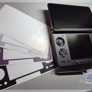 May include: Black Nintendo 3DS console with a stylus and several skin decals in various colors. The console is open, displaying two screens. The decals are white, gray, and black, with cutouts for the console's buttons and features.