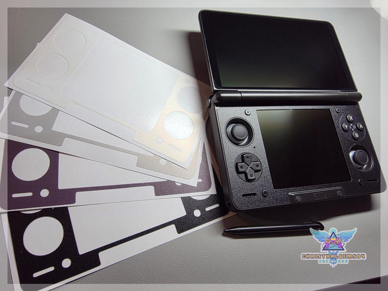 May include: Black Nintendo 3DS console with a stylus and several skin decals in various colors. The console is open, displaying two screens. The decals are white, gray, and black, with cutouts for the console's buttons and features.