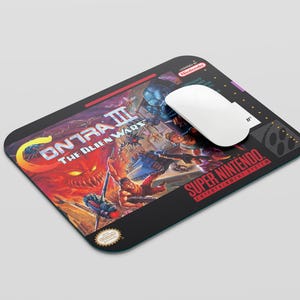 May include: A black mousepad with a retro video game design featuring the text "Contra III: The Alien Wars" and the Super Nintendo Entertainment System logo. The mousepad has a red, white, and blue color scheme.