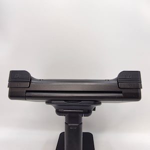 May include: A black gaming controller accessory mounted on a stand. The top edge features L1, L2, R1, and R2 shoulder buttons clearly labeled in embossed text. The device has a matte black finish and a horizontal rectangular shape with rounded corners.