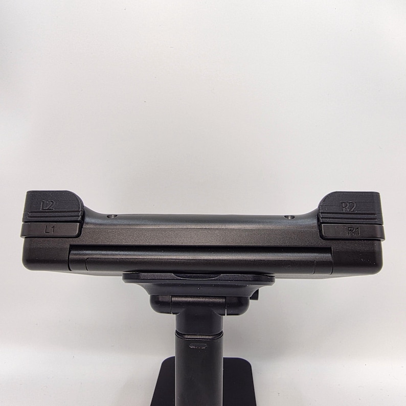May include: A black gaming controller accessory mounted on a stand. The top edge features L1, L2, R1, and R2 shoulder buttons clearly labeled in embossed text. The device has a matte black finish and a horizontal rectangular shape with rounded corners.