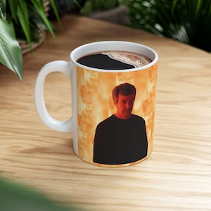 Pode incluir: Caneca de café de cerâmica branca com uma alça, apresentando um retrato de um homem com uma camisa preta contra um fundo laranja e amarelo. A caneca está cheia de líquido escuro, provavelmente café ou chá.