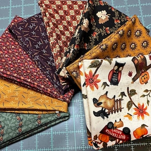 May include: A collection of folded fabric squares in various autumnal patterns. Designs include floral, geometric, and Halloween-themed prints with pumpkins, owls, and fall foliage. The fabrics are in shades of brown, orange, and green.
