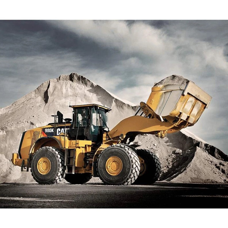 Puede incluir: Una cargadora de ruedas Caterpillar 980K amarilla con una gran cuchara levantada en el aire. La cargadora est&aacute; estacionada en un camino de tierra con un mont&oacute;n de tierra en el fondo.