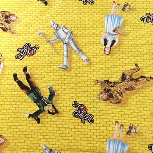 May include: Yellow fabric with a repeating pattern of characters from The Wizard of Oz. The characters include Dorothy, the Scarecrow, the Tin Man, and the Cowardly Lion. The fabric also features the words "The Wizard of Oz" repeated throughout.