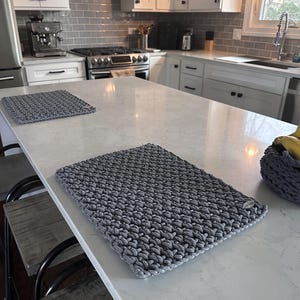 May include: Two gray crocheted placemats are displayed on a white countertop in a kitchen setting. The placemats have a textured, woven appearance. The kitchen features white cabinets, a stainless steel stove, and a coffee machine.
