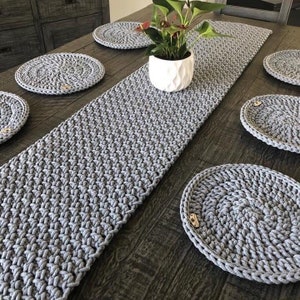 May include: A set of grey crocheted table linens consisting of a long rectangular table runner and five circular placemats. Each piece features a thick, textured knit pattern and a small, light brown wooden tag attached to the edge.