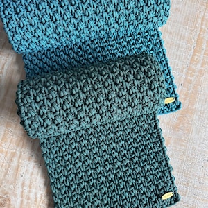 May include: Two rectangular crocheted mats featuring a textured, woven pattern in shades of teal and dark green. Each mat includes a small, oval-shaped gold-colored metal tag attached to the bottom corner with the text "CU Cords" engraved on it.