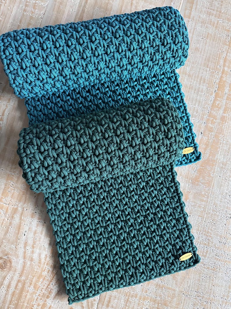 May include: Two rectangular crocheted mats featuring a textured, woven pattern in shades of teal and dark green. Each mat includes a small, oval-shaped gold-colored metal tag attached to the bottom corner with the text "CU Cords" engraved on it.
