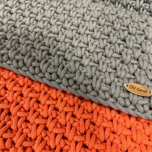 May include: Two layers of thick, hand-crocheted cord fabric in solid gray and bright orange. The gray layer sits atop the orange layer, both featuring a dense, interlocking weave pattern. An oval-shaped metallic tag engraved with the text OV Cords is attached to the corner of each piece.