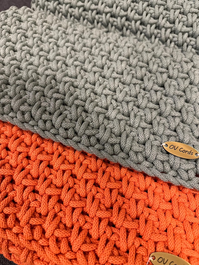 May include: Two layers of thick, hand-crocheted cord fabric in solid gray and bright orange. The gray layer sits atop the orange layer, both featuring a dense, interlocking weave pattern. An oval-shaped metallic tag engraved with the text OV Cords is attached to the corner of each piece.