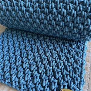 May include: A teal blue crocheted textile featuring a dense, textured stitch pattern. The fabric is folded over itself, creating a layered appearance. A small, oval-shaped gold-toned metal tag is attached to the corner, engraved with the text OV Cords.