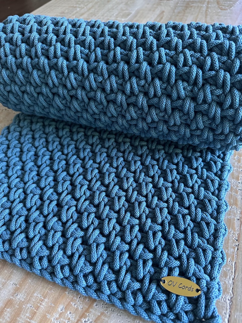 May include: A teal blue crocheted textile featuring a dense, textured stitch pattern. The fabric is folded over itself, creating a layered appearance. A small, oval-shaped gold-toned metal tag is attached to the corner, engraved with the text OV Cords.