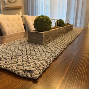 May include: A rectangular gray crocheted table runner with a textured, knotted stitch pattern. The runner features a small, oval-shaped metallic brand tag attached to one corner. It is laid flat on a dark brown wooden table surface.