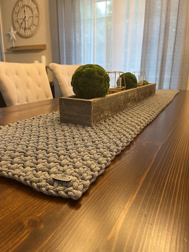 May include: A rectangular gray crocheted table runner with a textured, knotted stitch pattern. The runner features a small, oval-shaped metallic brand tag attached to one corner. It is laid flat on a dark brown wooden table surface.