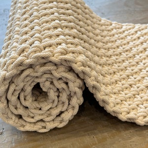 May include: A cream-colored textile runner featuring a thick, chunky knit construction with a repeating interlocking loop pattern. The fabric is rolled into a cylindrical shape at one end, revealing the dense, textured weave of the material.
