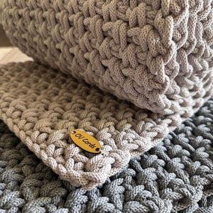 May include: A hand-crocheted textile item featuring a thick, textured cord weave in light beige and dark gray. The light beige section includes an oval-shaped wooden tag with the text OV Cords engraved in black, attached to the corner of the fabric.