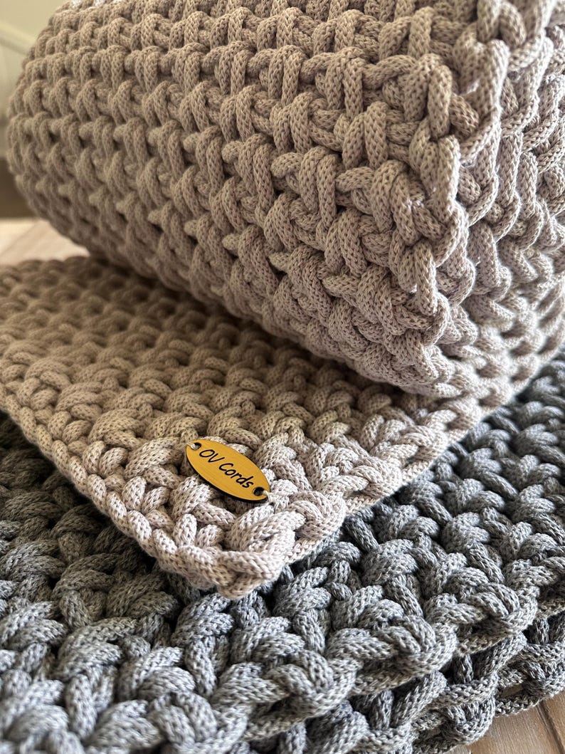 May include: A hand-crocheted textile item featuring a thick, textured cord weave in light beige and dark gray. The light beige section includes an oval-shaped wooden tag with the text OV Cords engraved in black, attached to the corner of the fabric.