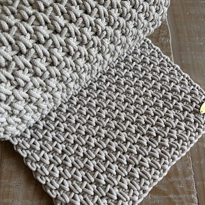 May include: A light beige textile item featuring a dense, hand-knotted macrame pattern with a thick cord construction. The piece is folded over itself, displaying a consistent, repeating woven texture throughout and a small gold-toned metal tag.