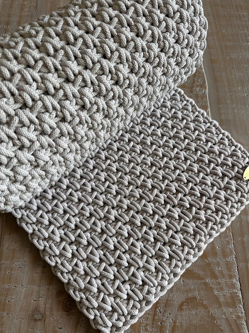 May include: A light beige textile item featuring a dense, hand-knotted macrame pattern with a thick cord construction. The piece is folded over itself, displaying a consistent, repeating woven texture throughout and a small gold-toned metal tag.