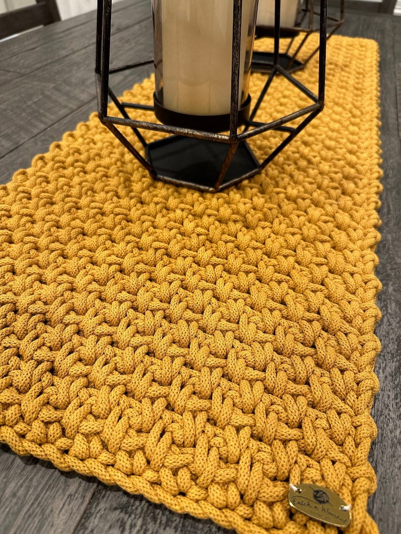 May include: A mustard yellow hand-knitted table runner with a thick, textured weave pattern. The runner features a small, oval-shaped metallic tag in the bottom right corner with the text "Patch a Moment" engraved on its surface, placed under a candle holder.