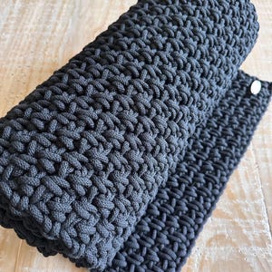 May include: A dark charcoal gray rectangular mat featuring a dense, hand-crocheted cord texture. The thick, woven material is rolled at one end, revealing a consistent, interlocking stitch pattern throughout the surface and a small, circular silver-toned metal tag.