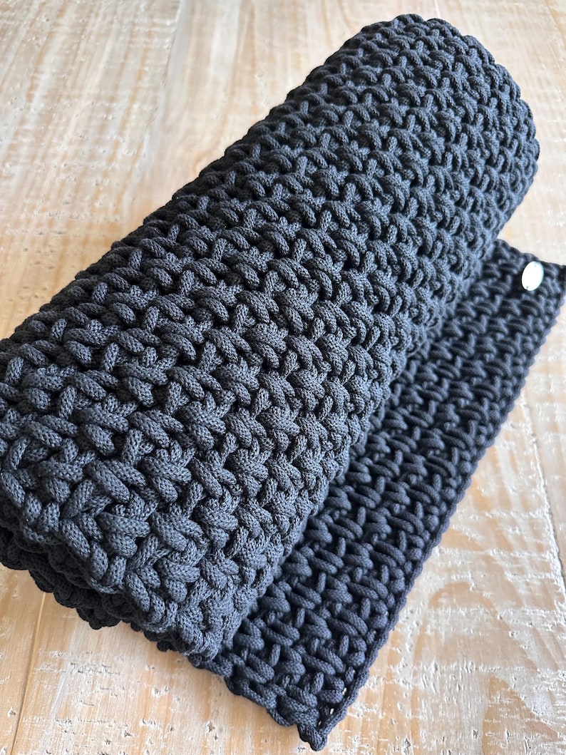 May include: A dark charcoal gray rectangular mat featuring a dense, hand-crocheted cord texture. The thick, woven material is rolled at one end, revealing a consistent, interlocking stitch pattern throughout the surface and a small, circular silver-toned metal tag.