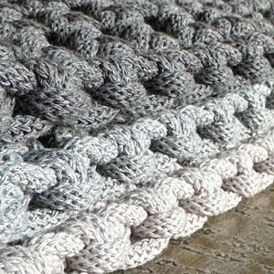 May include: A thick, hand-crocheted textile featuring a dense, raised stitch pattern. The piece displays a gradient color transition, shifting from a deep charcoal gray at the top to a lighter, soft silver-gray along the bottom edge.