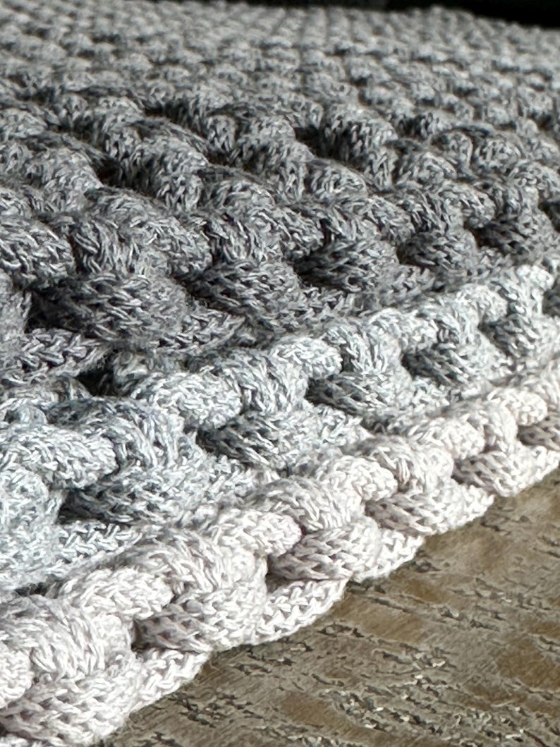 May include: A thick, hand-crocheted textile featuring a dense, raised stitch pattern. The piece displays a gradient color transition, shifting from a deep charcoal gray at the top to a lighter, soft silver-gray along the bottom edge.