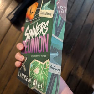 May include: A paperback book titled Sinners Reunion, a Slaycation Novel by Lauren Biel. The cover features a green and teal color scheme with road-themed graphics, including a map, a road sign reading Scenic DIEway, and a stop sign.
