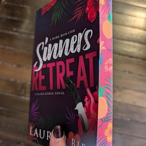 Puede incluir: Una portada de libro con un diseño tropical que presenta un cuchillo y un corazón. El título del libro es "Sinners Retreat" y se describe como "A Slaycation Novel". El nombre del autor es "Laur Biel".